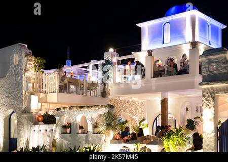 Eine griechische Taverne, in der Gäste an einem warmen Sommerabend an Tischen im Freien speisen. Das Restaurant ist mit blau-weißer Beleuchtung beleuchtet Stockfoto