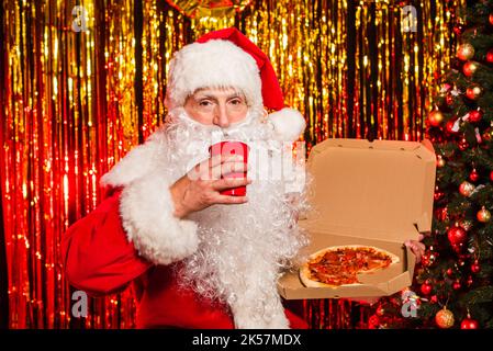 weihnachtsmann im Kostüm hält Plastikbecher und Pizza zum Mitnehmen in der Nähe von weihnachtsbaum und Lametta, Stock Bild Stockfoto