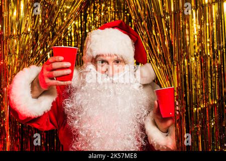 weihnachtsmann in Kostüm hält Plastikbecher und Blick auf Kamera in der Nähe Lametta, Stock Bild Stockfoto