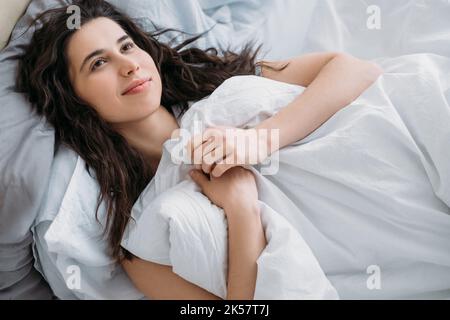 Gemütlicher Morgen. Wochenend-Freizeit. Heimhygge. Beauty-Wohlbefinden. Nachdenklich entspannt Tagträumerin Brünette Frau genießen im weißen gemütlichen Bett liegen lächelnd. Stockfoto