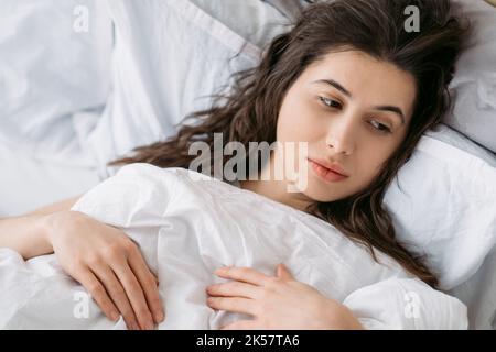 Trauriger Morgen. Am Wochenende melmütig. Schlaflosigkeit Problem. Nachdenklich enttäuscht verärgert Brünette Frau im Bett allein mit weißer Decke liegen. Stockfoto