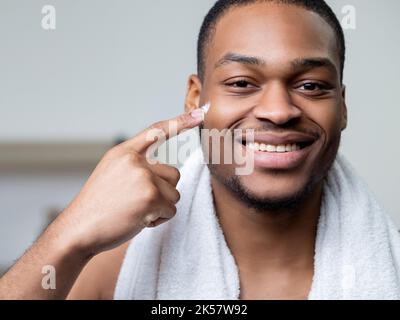 Männliche Hautpflege. Gesichtsbehandlung. Werbung für kosmetische Produkte. Selbstbewusster, fröhlicher afrikanischer Mann mit weißem Handtuch, das zeigt, dass er mit Fingercreme auf d zeigt Stockfoto