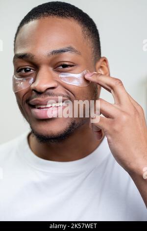 Gesichtsbehandlung für Männer. Hautverjüngung. Werbung für kosmetische Produkte. Zufriedene Afrikaner in weißen T-Shirt Anwendung Kollagen Gel Gesicht Augenflecken Smilie Stockfoto