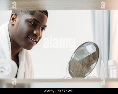 Morgendliche Routine im Bad. Lächelnder schwarzer Mann. Perfekte Haut. Hygiene-Pflege. Glücklich zuversichtlich afrikanischen Kerl weißes Handtuch auf der Schulter bewundernde Reflexion ich Stockfoto