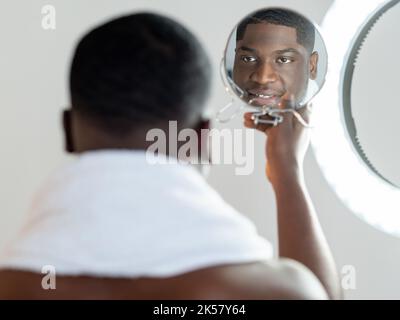 Morgenpflege. Schwarzer Hipster. Gesichtspflege. Perfekte Haut. Tägliche Routine. Glücklich zuversichtlich afrikanischen Kerl weißes Handtuch auf der Schulter bewundernd reflektieren Stockfoto