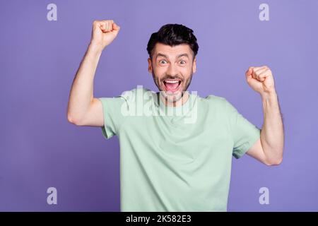 Portrait von fröhlich beeindruckt aufgeregt Mann mit Brunet Frisur gekleidet T-Shirt ekstatisch heben Fäuste isoliert auf lila Hintergrund Stockfoto