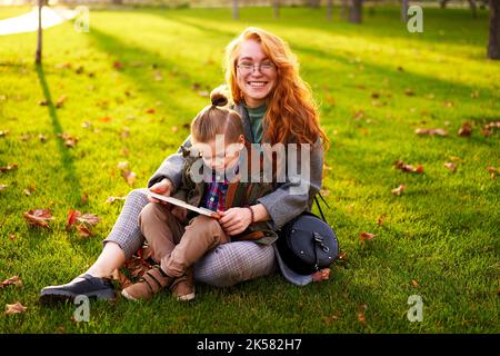 Rotschopf Frau liest Buch mit kleinen Jungen auf Gras im Stadtpark sitzen. Die junge Mutter unterrichtet ihren Sohn mit der ersten Klasse und macht Hausaufgaben am sonnigen Herbsttag Stockfoto