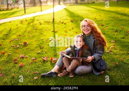 Rotschopf Frau liest Buch mit kleinen Jungen auf Gras im Stadtpark sitzen. Die junge Mutter unterrichtet ihren Sohn mit der ersten Klasse und macht Hausaufgaben am sonnigen Herbsttag Stockfoto