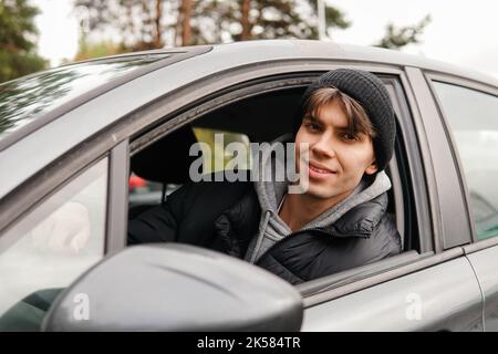 Junger lächelnder Student, der in seinem neuen Auto sitzt. Mieten Sie ein Auto oder kaufen Sie das erste Auto Konzept. Stockfoto