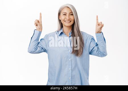 Porträt einer lächelnden, gesunden asiatischen älteren Frau, 50 Jahre alte Dame, die die Finger nach oben zeigt, Werbung zeigt, nach oben, über dem weißen Studio stehend Stockfoto