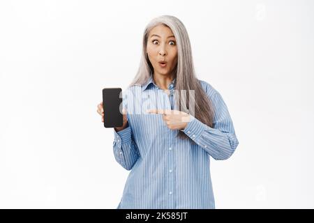 Aufgeregt Senior Lady, asiatische Großmutter zeigt Handy-Bildschirm, zeigt auf Smartphone-App, stand auf weißem Hintergrund Stockfoto