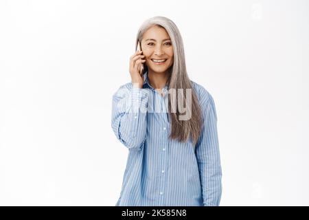 Porträt einer glücklich lächelnden asiatischen älteren Frau, einer alten Dame, die Smartphone benutzt, auf dem Mobiltelefon spricht und mit dem Mobiltelefon vor weißem Hintergrund steht Stockfoto