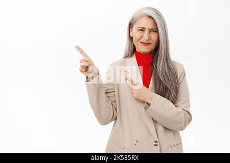 Asiatische Geschäftsfrau, Unternehmerin zeigt mit enttäuschtem Gesicht nach links und steht aufgeregt vor weißem Hintergrund Stockfoto
