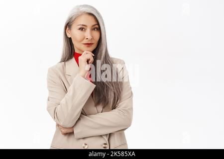 Bild einer eleganten asiatischen Senior-Frau im Anzug, lächelnd und fasziniert, nachdenklicher Gesichtsausdruck, der auf weißem Hintergrund steht Stockfoto