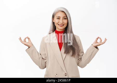 Behaltet die Ruhe. Asiatische ältere Frau im Business-Anzug, meditierend, tief durchatmen, Luft einatmen und sich entspannen, stehend über weißem Hintergrund Stockfoto