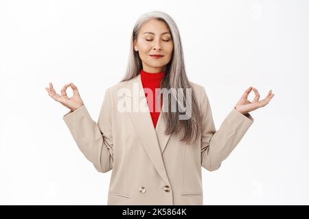 Behaltet die Ruhe. Asiatische ältere Frau im Business-Anzug, meditierend, tief durchatmen, Luft einatmen und sich entspannen, stehend über weißem Hintergrund Stockfoto