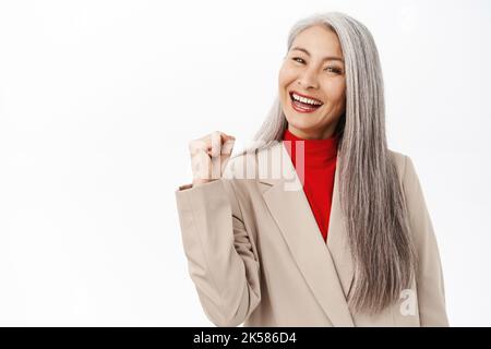 Ja, erfolgreich. Lächelnde asiatische ältere Frau, Bürodame mittleren Alters, die Erfolge feiert, zufrieden aussiegt und auf weißem Hintergrund steht Stockfoto