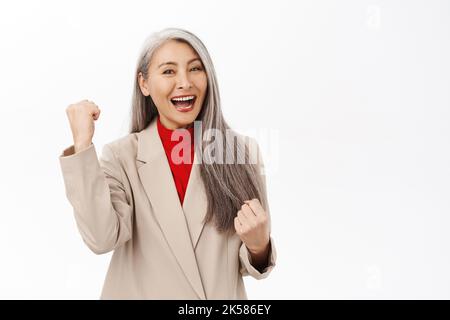 Glückliche asiatische Frau mittleren Alters erreichen Ziel und feiern, machen Faust Pumpe, sagen ja und lächeln zufrieden, zufrieden mit smth, über weiß stehen Stockfoto