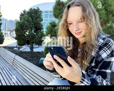 IPhone 13 Teen Redhead Mädchen auf städtischen Straße Wolkenkratzer Hintergrund trägt Kopfhörer mit Smartphone Musik hören Hörbuch Reiseführer Podcast während des Spazierens in der Stadt Kanada 09,2022 Stockfoto