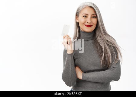 Selbstbewusst lächelnde asiatische Senior-Frau, schöne koreanische weibliche Modell mit Kreditkarte mit zufrieden, entspannten Gesichtsausdruck, über weiß stehend Stockfoto
