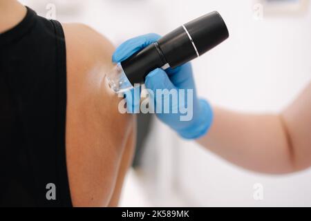 Ärztin und weibliche Klientin in dermatologischer Klinik. Makrofoto des Dermatologen verwendet Dermatoskop für die Gesichtshautuntersuchung. Stockfoto