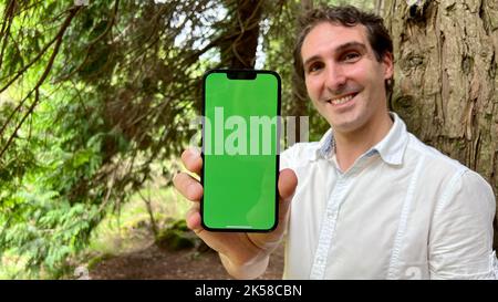 Sehr hübscher Mann französisch italien nicht hält ein Telefon mit einem grünen Hahn im Wald in einem weißen Hemd stehen und lächeln zeigt einen Finger auf dem Bildschirm kann für die Werbung verwenden. Hochwertige 4K-Aufnahmen Stockfoto