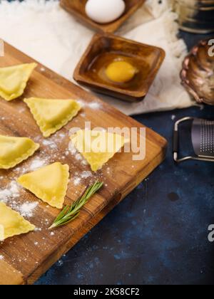 Nahaufnahme. Rohe Ravioli, Küchenutensilien und Zutaten auf einem Holzschneidebrett auf dunkelblauem Hintergrund. Italienische Küche Rezepte, der Prozess der m Stockfoto