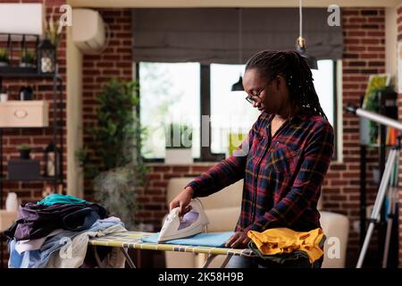afroamerikanische Frau bügeln Kleidung zu Hause, Wäsche-und Bügelservice Tag, unabhängigen Lebensstil in städtischen Wohnung, moderne Hausfrau tun Aufgaben und Hausarbeit zu Hause. Vorderansicht. Stockfoto