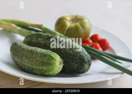 Weißer Teller mit frischem Gemüse auf Holztisch. Zwei Gurken, rote Kirschtomaten, grüne rissige Tomate mit vertikalen Spalten, grüner Schnittlauch. Gesund Stockfoto