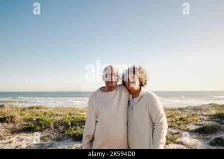 Porträt eines glücklichen älteren Paares, das lächelt an der Kamera, während es am Strand zusammen steht. Ein romantisches, älteres Paar, das einen entspannenden Urlaub am Meer hat Stockfoto