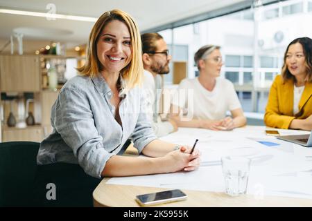 Fröhliche junge Designerin, die die Kamera anlächelt, während sie in einem Meeting mit ihrem Team sitzt. Gruppe von innovativen Geschäftsleuten, die kreative Ideen austauschen, während w Stockfoto
