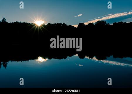 Die Sonne untergeht in einem glorreichen Starburst hinter silhouettierten Bäumen, die den Rand eines Sees säumen. Stockfoto