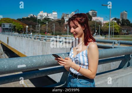 Junge Rotschopf-Frau, die Nachrichten auf ihrem Handy überprüft, lächelnde Einstellung, nachdenkliche Einstellung, zufriedene Haltung. Speicherplatz kopieren. Ausrichtung im Querformat. Stockfoto