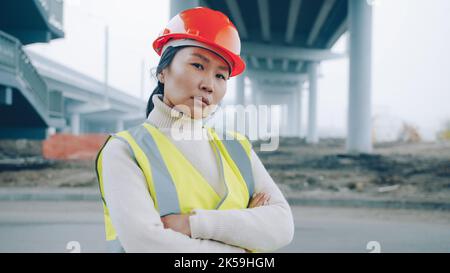 Porträt einer asiatischen Bauingenieurin, die mit gekreuzten Armen im Gebäudebereich steht und selbstbewusst bei der Arbeit auf die Kamera schaut Stockfoto