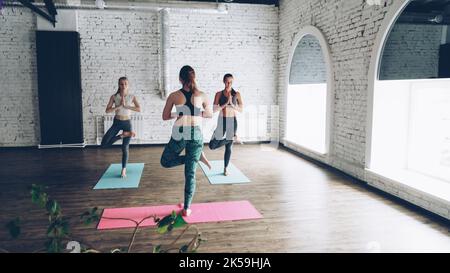 Eine erfahrene Yogalehrerin zeigt zwei attraktiven Schülerinnen beim Gruppentraining im Wellnesscenter eine ausgleichende Haltung. Weiße Wände, helle Matten, große Fenster sind sichtbar. Stockfoto