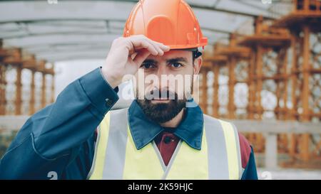 Fröhlicher Baumeister setzt sich Schutzhelm an und lächelt, steht allein auf der Baustelle und schaut in die Kamera. Schutz und einheitliches Konzept. Stockfoto