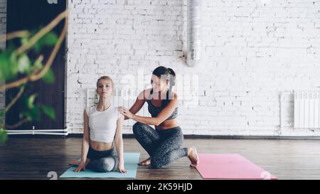 Fröhliche attraktive Yoga-Lehrer hilft neuen Schüler Master Kuh Gesicht Pose während eins-zu-eins-Klasse in modernen Sportzentrum. Frauen lächeln und lachen, freundliche Atmosphäre. Stockfoto