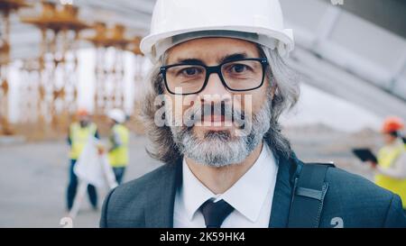 Nahaufnahme des reifen Architekten, der vor der Baustelle mit Helm steht und die Kamera anschaut. Projekte und Gebäudekonzept. Stockfoto