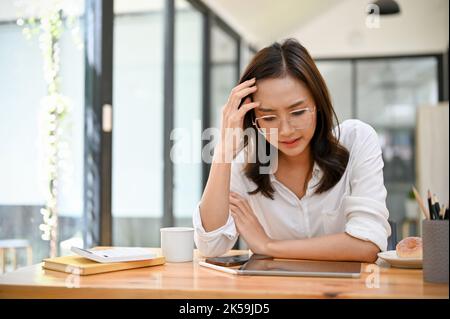 Eine gestresste und ernsthafte, tausendjährige asiatische Geschäftsfrau oder Managerin, die an Depressionen und Kopfschmerzen leidet, sitzt an ihrem Schreibtisch. Stockfoto