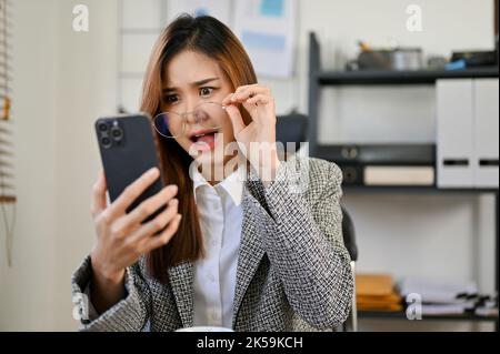 Schockiert und erstaunt berührte die junge asiatische Geschäftsfrau ihre Brille, schaute auf ihren Smartphone-Bildschirm und las unerwartete Nachrichten oder E-Mails. Stockfoto