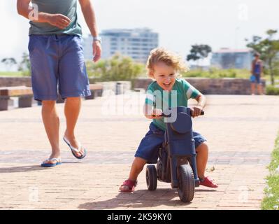 HES geboren, um ein Biker-Junge zu sein. Ein glücklicher Kleinkind Junge Reiten ist Spielzeug Motorrad draußen, während sein Vater hinter ihm läuft. Stockfoto