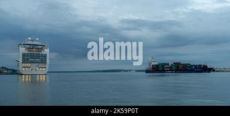 Cobh, Irland - 15. August 2022: Panoramablick auf ein großes Kreuzschiff auf dem Pier in Cobh mit einem großen Frachtschiff, das in der Dämmerung in den Hafen von Cork einfährt Stockfoto