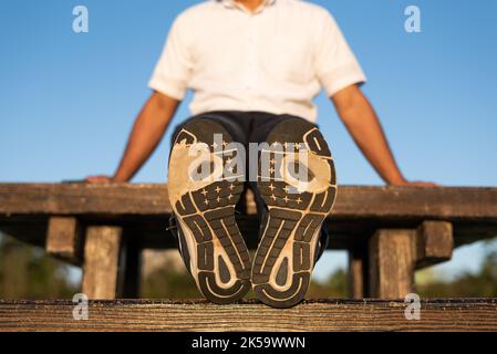 Ein Mann, der Laufschuhe mit abgenutzter Außensohle trägt, sitzt auf einem Tisch auf der Bank Stockfoto