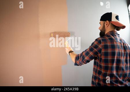 Hübscher junger bärtiger Maler in Baseballmütze und kariertem Hemd, der an der Wand mit Farbrolle malte. Mann Malerei Haus in DIY Stockfoto