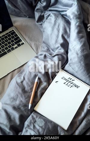 Sparsame Tipps für das Leben, Sparkonzept mit Einkaufsliste im Notebook in der Nähe von Laptop und Tee-Kaffeetasse in Bad zu Hause Stockfoto