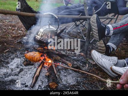 Junge und fröhliche Freunde sitzen am Lagerfeuer, kochen den Wasserkocher und braten Brot in der Natur. Kochen am Lagerfeuer Stockfoto