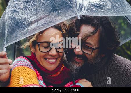 Liebe zwischen erwachsenen Mann und Frau, die gemeinsam unter einem transparenten Regenschirm Spaß haben, an regnerischen Tagen bei Freizeitaktivitäten im Freien. Glückliches Paar in Beziehung Stockfoto