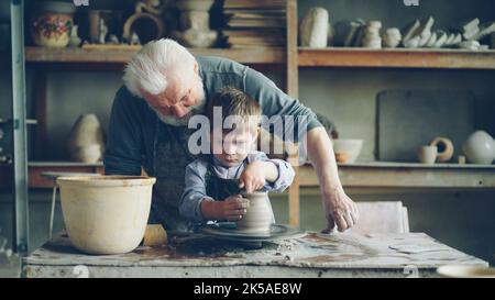 Konzentrierter Junge formt Ton in Keramikvase auf drehendes Wurfrad und sein liebevoller erfahrener Großvater hilft ihm. Töpferei und Familientradition Konzept. Stockfoto