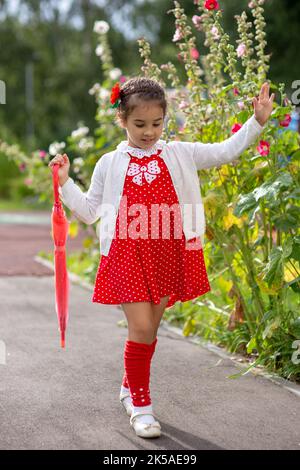 Ein niedliches kleines Mädchen geht an einem Sommertag in einem roten Kleid und einer weißen Bluse mit einem rosa gefalteten Regenschirm spazieren Stockfoto