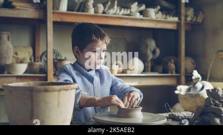 Nettes neugieriges Kind macht Tontopf auf drehendes Wurfrad in der professionellen Werkstatt des Großvaters. Braune Keramikvasen, handgefertigte Töpfe, Töpferwerkzeuge sind sichtbar. Stockfoto